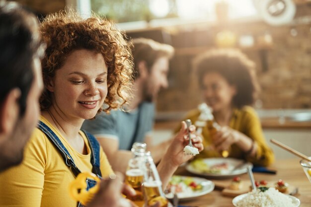 家で友達と昼食をとりながらビール瓶で乾杯する若いカップル。赤毛の女性に焦点を当てています。