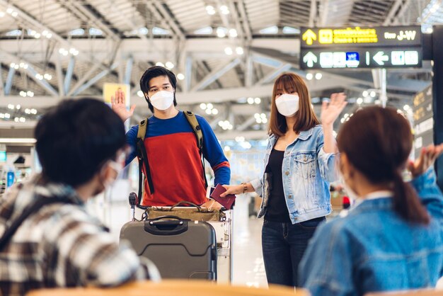 国際線​ターミナル​空港​で​の​長い​旅行​休暇​の​フライト​の​後​、​こんにちは​を​振って​友人​と​挨拶する​サージカルマスク​の​顔​の​保護​を​身​に​着けている​コロナ​ウイルス​の​検疫中​の​若い​カップル​の​旅行者