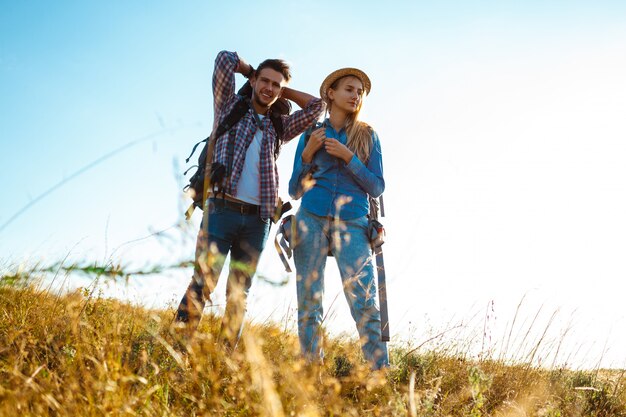 Young couple of travelers with backpacks smiling, standing in field