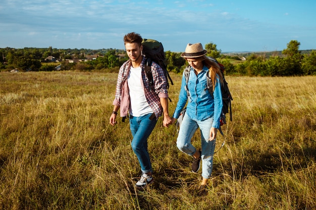 Young couple of travelers with backpacks smiling, walking in field