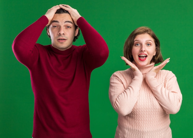 Young couple on valentine's day concerned man keeping hands on head excited woman showing empty hands under chin both looking at front isolated on green wall