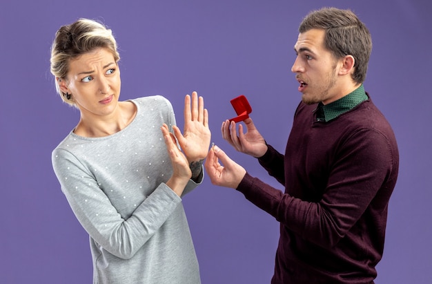 Young couple on valentines day confused guy giving wedding ring to unpleased girl isolated on blue background