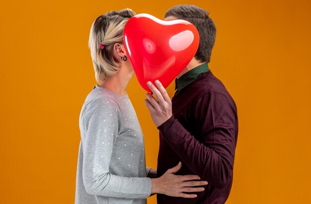 발렌타인 데이에 젊은 부부는 주황색 배경에 고립 된 심장 풍선으로 얼굴을 덮었습니다.