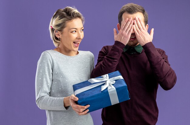 Молодая пара в день святого валентина взволнованная девушка дарит подарочную коробку парню, изолированному на синем фоне