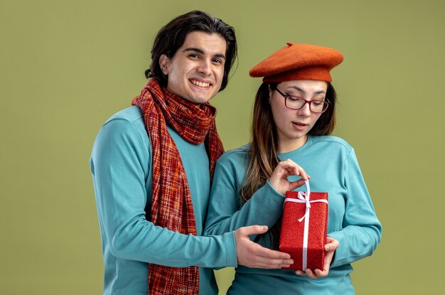 Young couple on valentines day guy wearing scarf girl wearing hat girl holding gift box isolated on olive green background