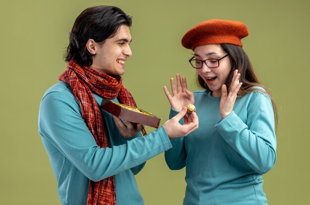 Young couple on valentines day guy wearing scarf girl wearing hat smiling guy giving box of candies isolated on olive green background