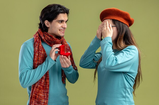 Young couple on valentines day guy wearing scarf girl wearing hat smiling guy giving wedding ring to girl isolated on olive green background