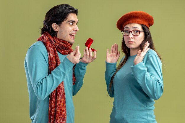 Young couple on valentines day guy wearing scarf girl wearing hat smiling guy giving wedding ring to girl isolated on olive green background