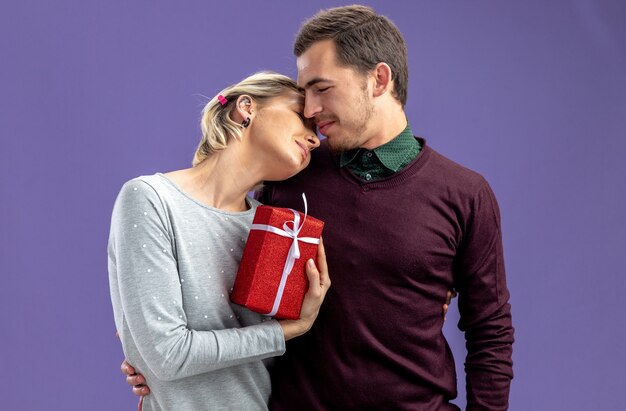 Young couple on valentines day hugged each other girl holding gift box isolated on blue background