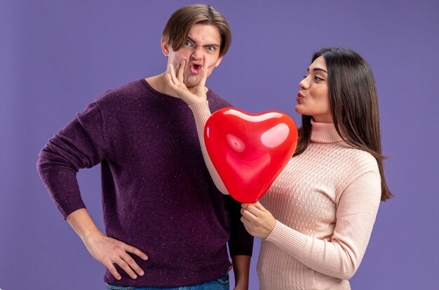 Young couple on valentines day pleased girl holding heart balloon grabbed guy chin isolated on blue background