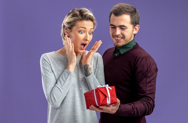 Young couple on valentines day pleased guy giving gift box to excited girl isolated on blue background