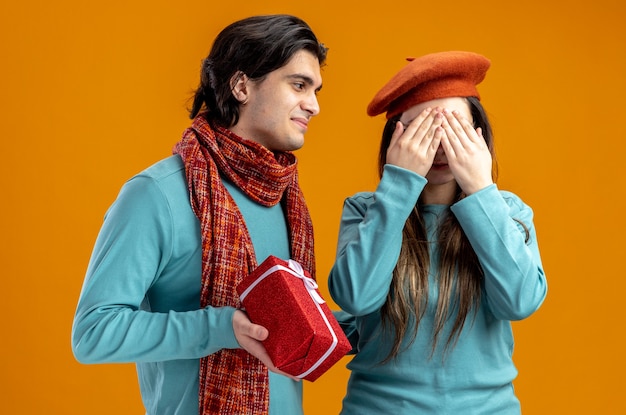 Young couple on valentines day pleased guy giving gift box to girl isolated on orange background