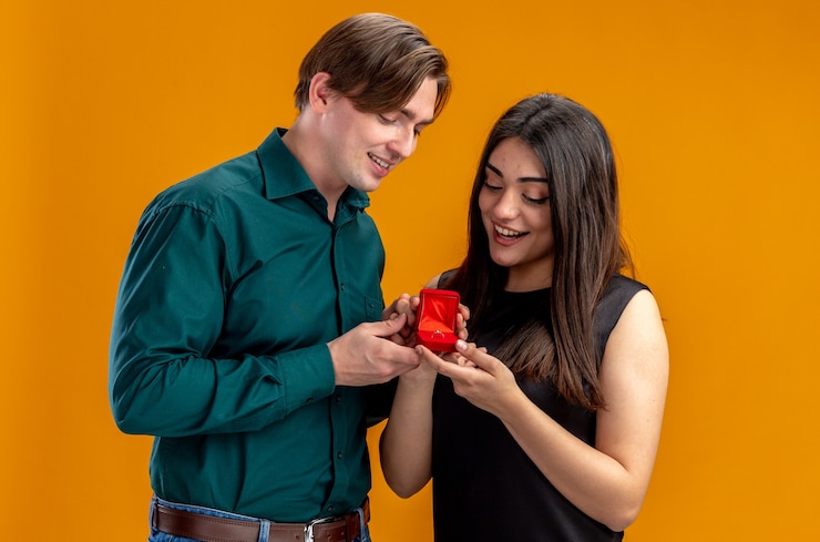 मुफ़्त फोटो young couple on valentines day pleased guy giving wedding ring to smiling girl isolated on orange background
