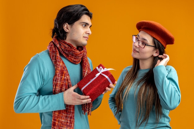 Young couple on valentines day pleased guy wearing scarf holding gift box looking at each other isolated on orange background