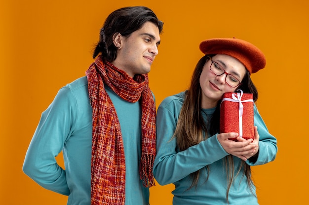 バレンタインデーの若いカップルは、オレンジ色の背景で隔離のギフトボックスを持つ女の子を見てスカーフを身に着けている男を喜ばせた