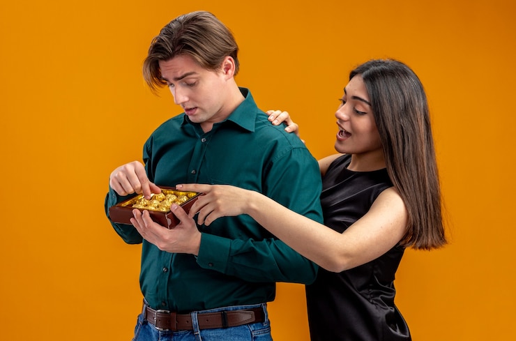 Young couple on valentines day smiling girl standing next to guy with box of canies isolated on orange background