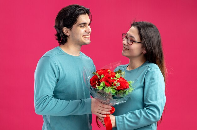 Young couple on valentines day smiling guy giving bouquet to pleased girl isolated on pink background