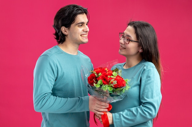 ピンクの背景で隔離の幸せな女の子に花束を与えるバレンタインデーの笑顔の若いカップル