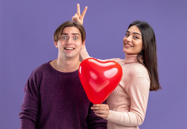 Young couple on valentines day smiling holding heart balloon giving bunny ears to guy isolated on blue background