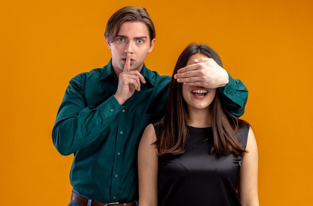 Young couple on valentines day strict guy covered girl eyes with hand showing silence gesture isolated on orange background