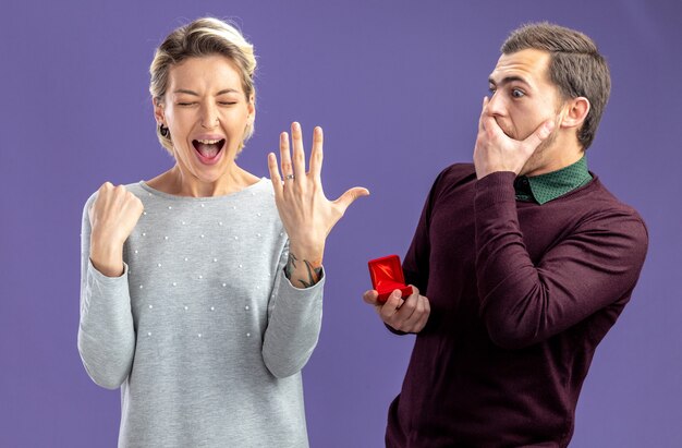 Young couple on valentines day surprised guy giving wedding ring to excited girl isolated on blue background