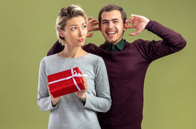 Free photo young couple on valentines day thinking girl holding gift box smiling guy standing behind girl closed ears isolated on olive green background