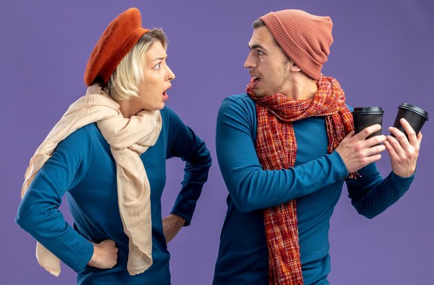 Young couple wearing hat with scarf on valentine's day angry girl looking at guy with cup of coffee isolated on blue background