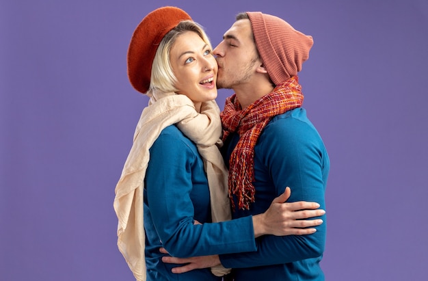 Young couple wearing hat with scarf on valentine's day pleased guy kissing girl cheek isolated on blue background