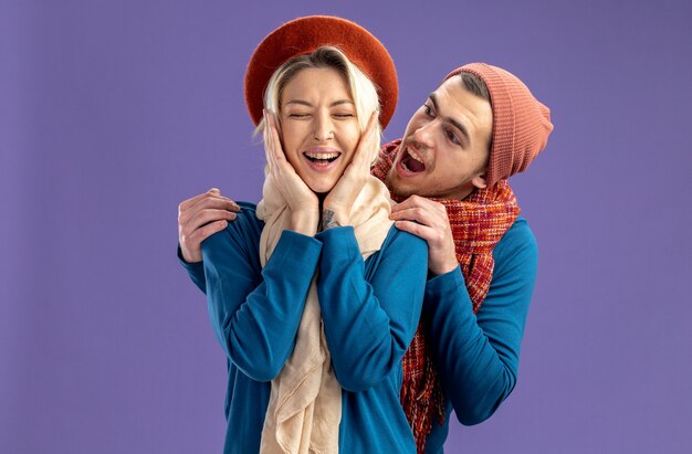 Young couple wearing hat with scarf on valentine's day pleased with closed eyes girl putting hands on cheeks guy standing behind girl isolated on blue background