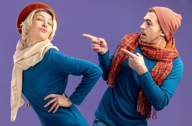 Young couple wearing hat with scarf on valentine's day surprised guy points at pleased girl isolated on blue background