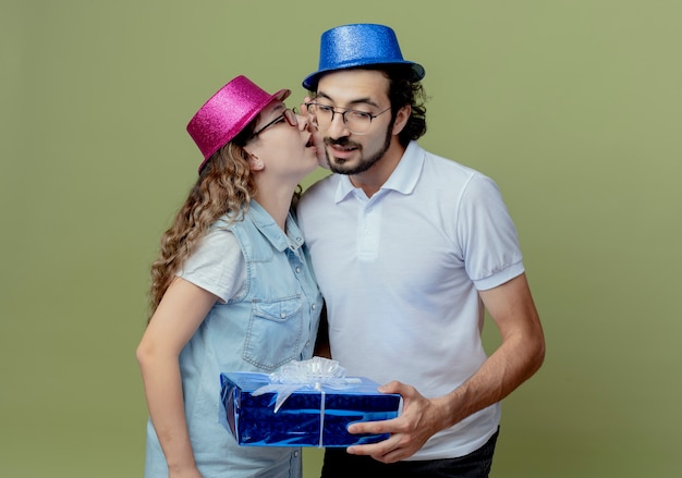 Young couple wearing pink and blue hat girl whisper on guy ear and guy holding gift box isolated on olive green