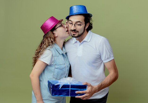 Молодая пара в розово-синей шляпе, девушка шепчет на ухо парню и парень, держащий подарочную коробку, изолированную на оливково-зеленом