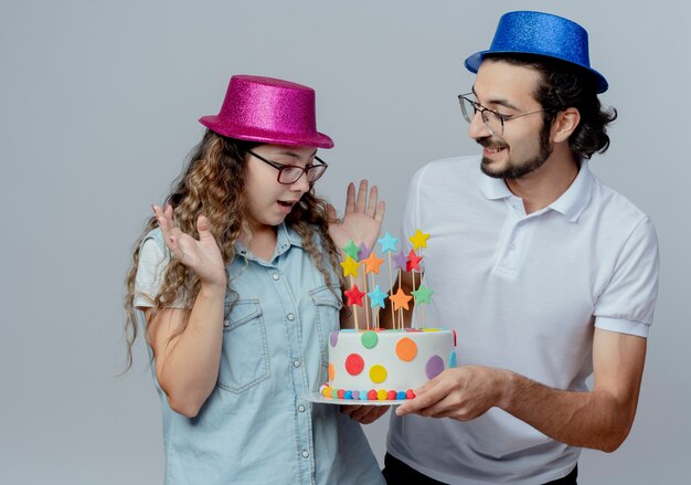 Giovane coppia che indossa cappello rosa e blu ragazzo soddisfatto dà la torta di compleanno alla ragazza sorpresa isolata su bianco