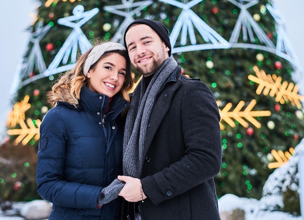 Young couple wearing standing near a city Christmas tree, enjoying spending time together