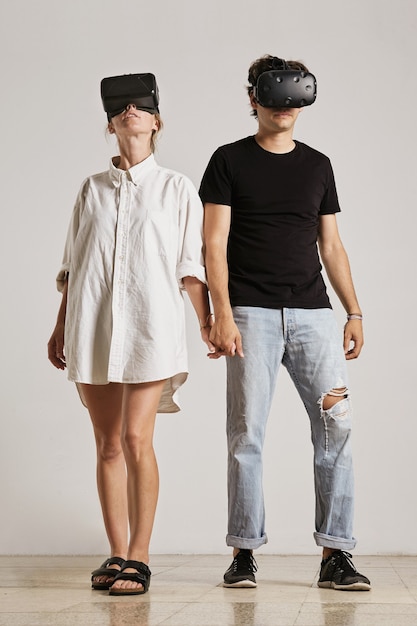 흰 벽과 나무 바닥이있는 방에서 서로 다른 방향으로보고 손을 잡고 VR 헤드셋을 착용 한 젊은 부부