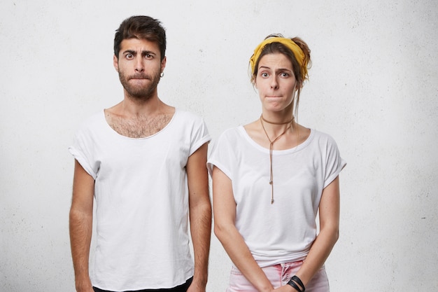 Young couple in white T-shirts standing close to each other pressing their lips with displeasure having bad mood