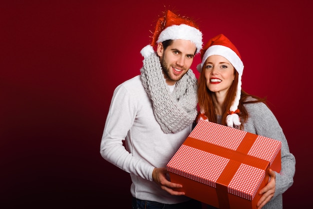 Young couple with a christmas gift dressed with winter clothes and santa hat on red background