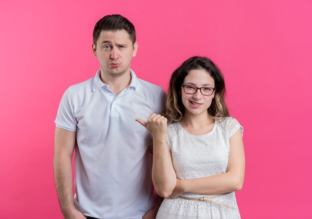 Young couple woman pointing with finger at her confused boyfriend standing over pink wall