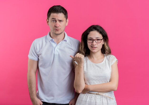 Young couple woman pointing with finger at her confused boyfriend standing over pink wall