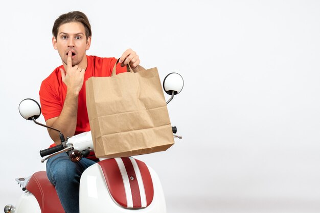 Young courier guy in red uniform sitting on scooter holding paper bag thinking deeply on white wall