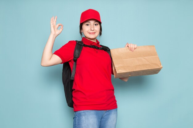 young courier in red polo red cap black backpack holding package smiling on blue