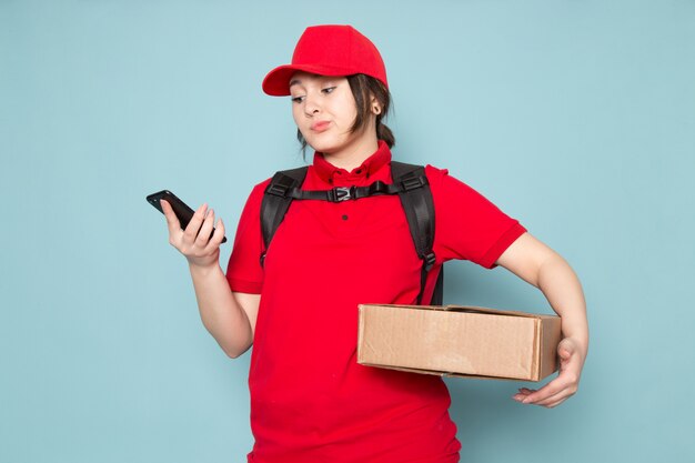 young courier in red polo red cap black backpack holding package using phone on blue