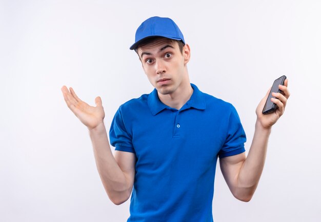 Молодой курьер в синей форме и синей кепке в замешательстве держит телефон