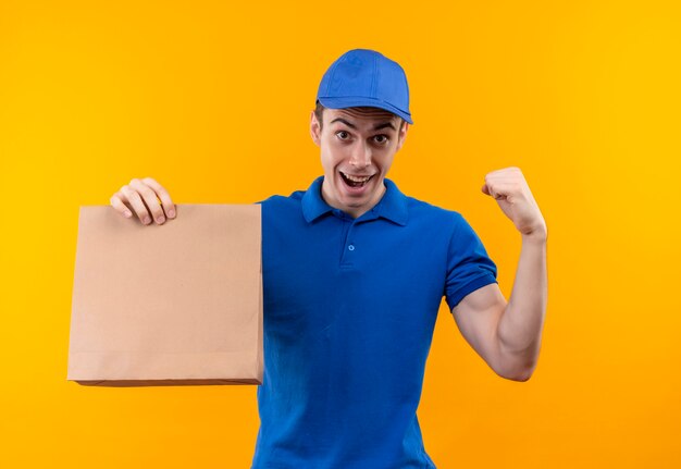 Young courier wearing blue uniform and blue cap doing happy fist holds a bag