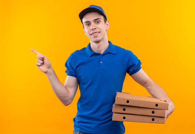 Young courier wearing blue uniform and blue cap doing happy forefinger to the right holds boxes