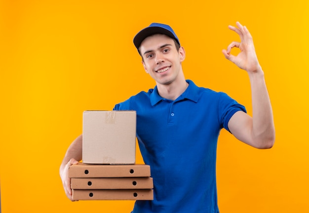 Young courier wearing blue uniform and blue cap doing happy ok with thumbs holds boxes