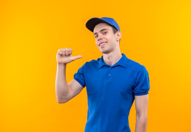 Young courier wearing blue uniform and blue cap doing happy thumbs on himself