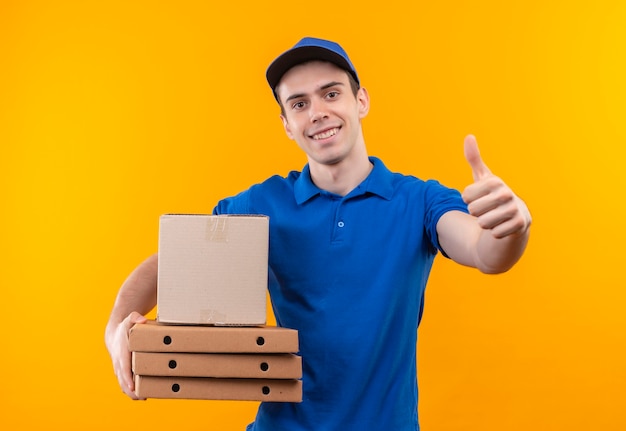 Young courier wearing blue uniform and blue cap doing happy thumbs up holds boxes