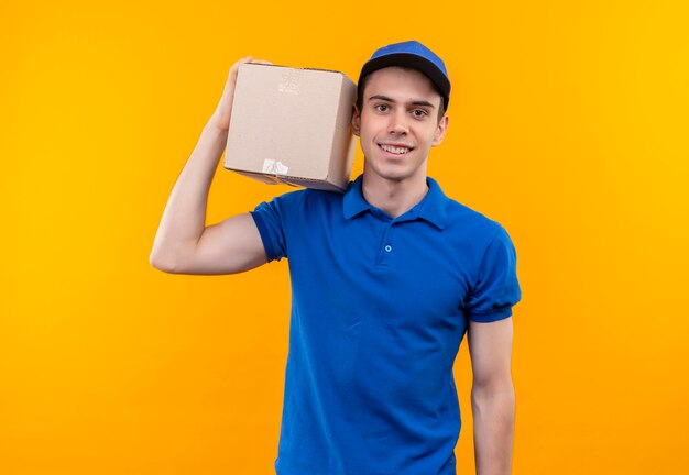 Young courier wearing blue uniform and blue cap happily holds a box on the right shoulder