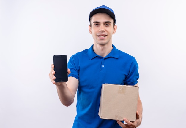 Young courier wearing blue uniform and blue cap happily holds a telephone and bag